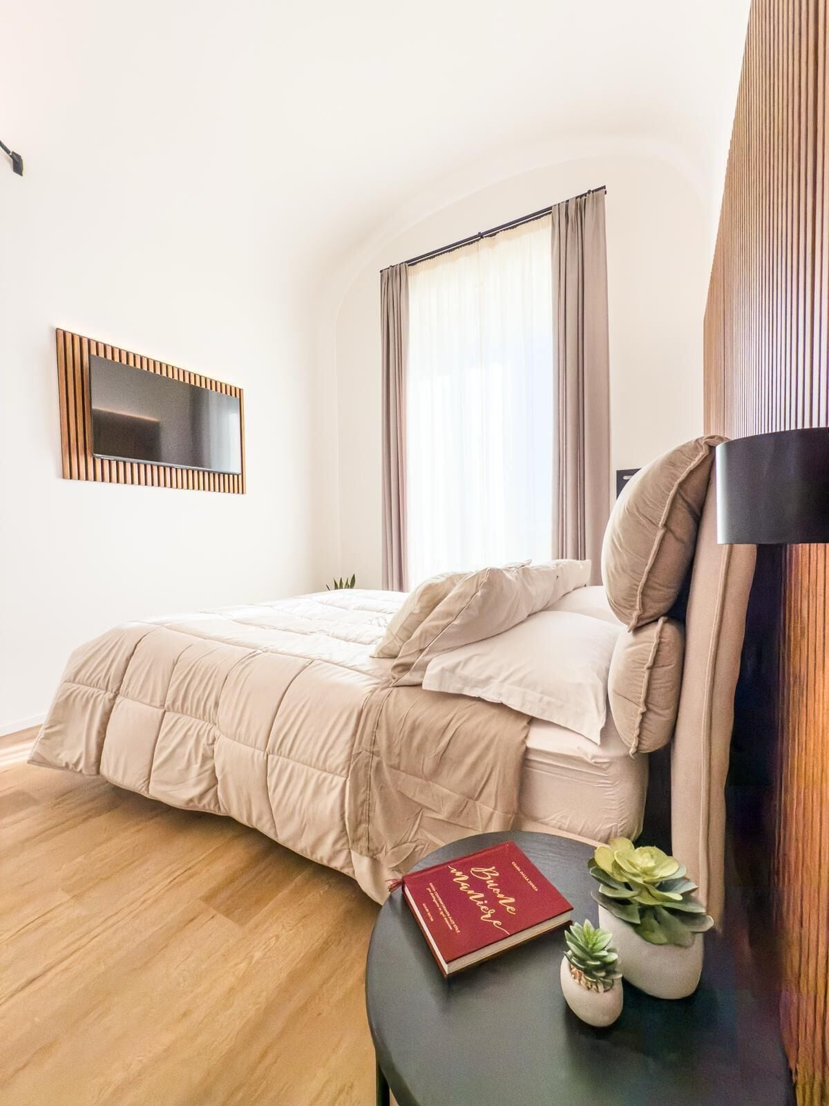 A neatly made bed with a beige comforter and pillows, a bedside table with a book and two small potted plants, and a large mirror on the wall.