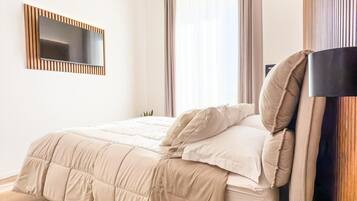 A neatly made bed with a beige comforter and pillows, a bedside table with a book and two small potted plants, and a large mirror on the wall.