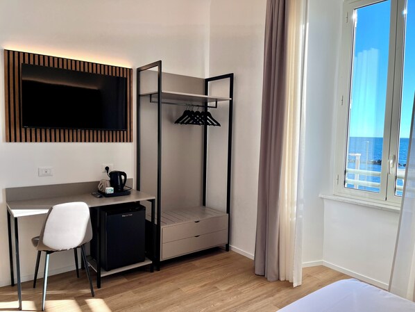 Quarto compacto com escrivaninha, cadeira e pequeno frigorífico. Uma cama beliche com escada articulada está encostada à parede, e uma janela oferece vista para o mar.