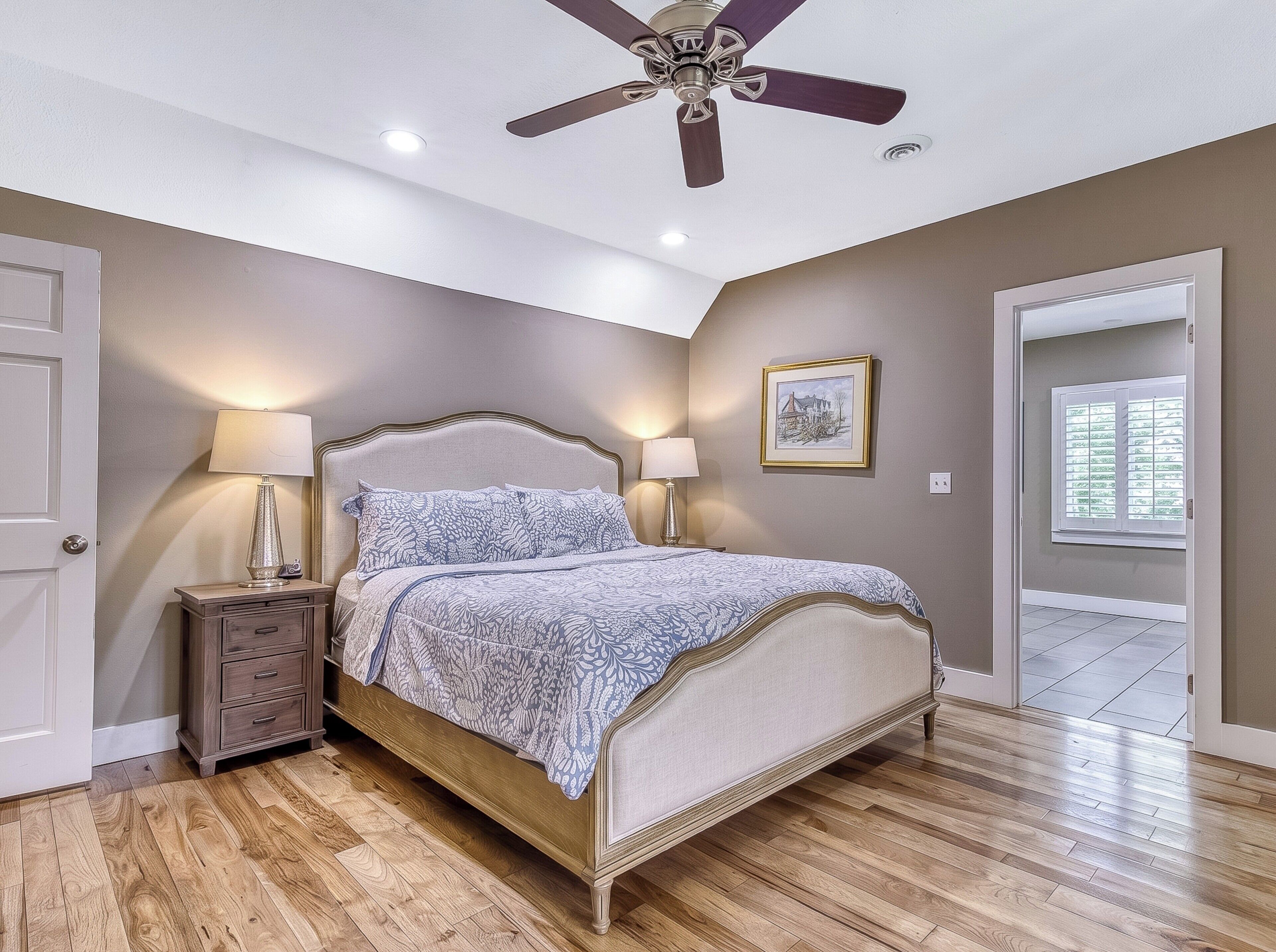 Une chambre à coucher comprenant un lit, deux tables de chevet, un ventilateur de plafond, une fenêtre avec des stores et une porte.