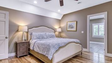 Une chambre à coucher comprenant un lit, deux tables de chevet, un ventilateur de plafond, une fenêtre avec des stores et une porte.