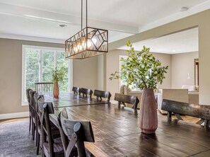 Comedor con gran mesa de madera, varias sillas, lámpara colgante y dos plantas en macetas.