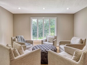 Una habitación con un gran ventanal, paredes color beige y una zona de estar con un otoman tapizado y dos sillones.