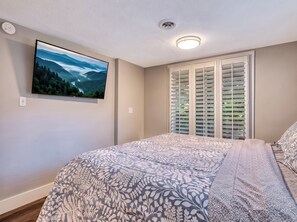 Ein Schlafzimmer mit einem Bett, einem Wand-Fernseher, einem Fenster mit Jalousien und einer Deckenleuchte.