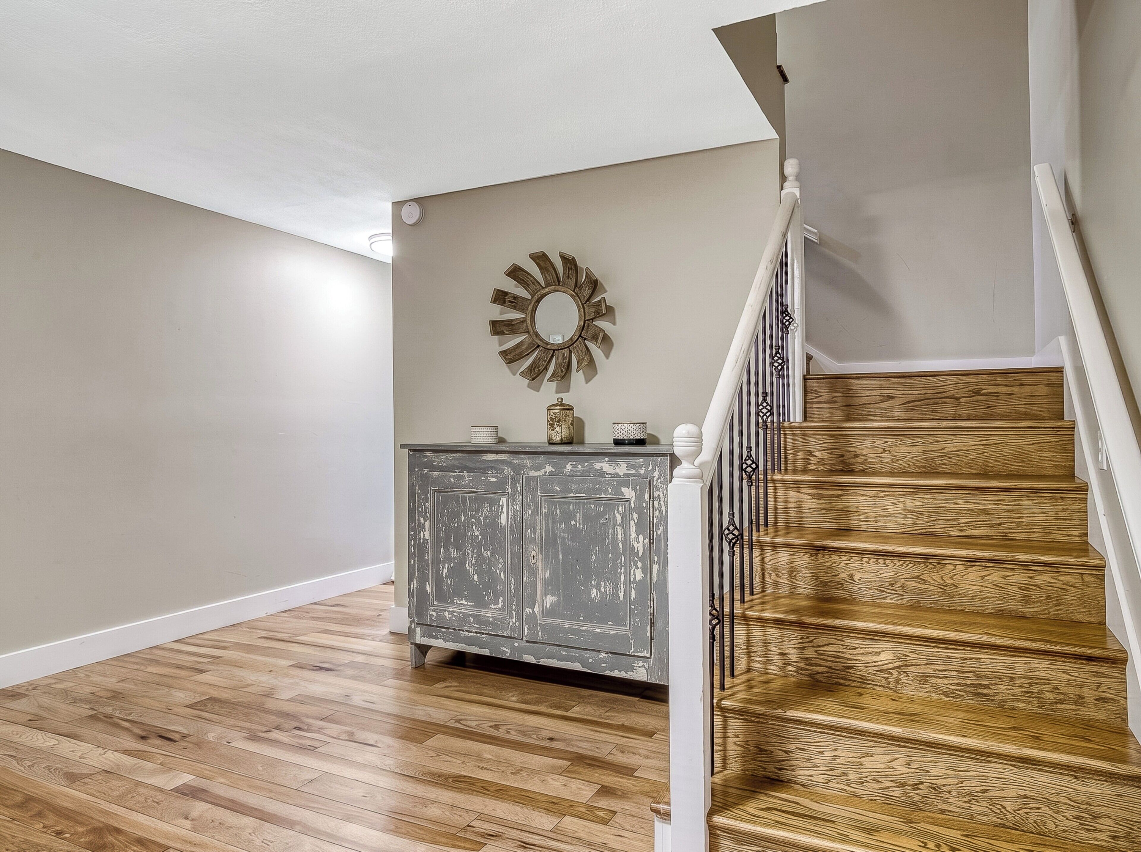 Una escalera de madera con peldaños que lleva a una plataforma, una barandilla blanca y un gabinete gris con un espejo decorativo encima.
