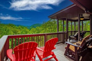 Eine Holzterrasse mit roten Adirondack-Stühlen und einer Korbsitzgruppe, mit Blick auf eine bewaldete Landschaft.