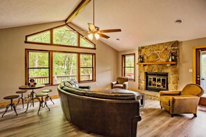 A living room with a stone fireplace, large windows, a ceiling fan, and a cozy seating area. - Big Bear Lodge | Sleeps 28 | Mtn Views + Hot Tub (Sevierville)