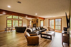 A spacious living room with a large leather sofa, a coffee table, and a fireplace. There are large windows providing natural light and views of the outdoors. - Big Bear Lodge | Sleeps 28 | Mtn Views + Hot Tub (Sevierville)