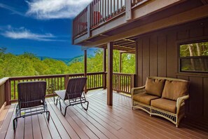 A wooden deck with a balcony, wicker chairs, a sofa, and a view of a forested landscape. - Big Bear Lodge | Sleeps 28 | Mtn Views + Hot Tub (Sevierville)