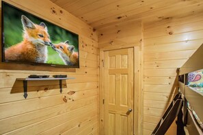 Interior de una cabaña de madera con un televisor en la pared, una puerta y una litera con una manta colorida.