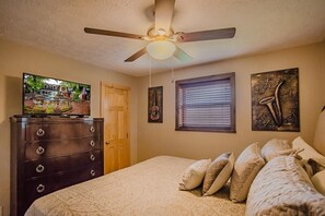 Une chambre à coucher comprenant un lit, une commode, un ventilateur de plafond, un téléviseur et des œuvres d’art accrochées aux murs.