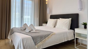 A modern bedroom with a large bed, white bedding, a dark headboard, and a bedside table with a plant.