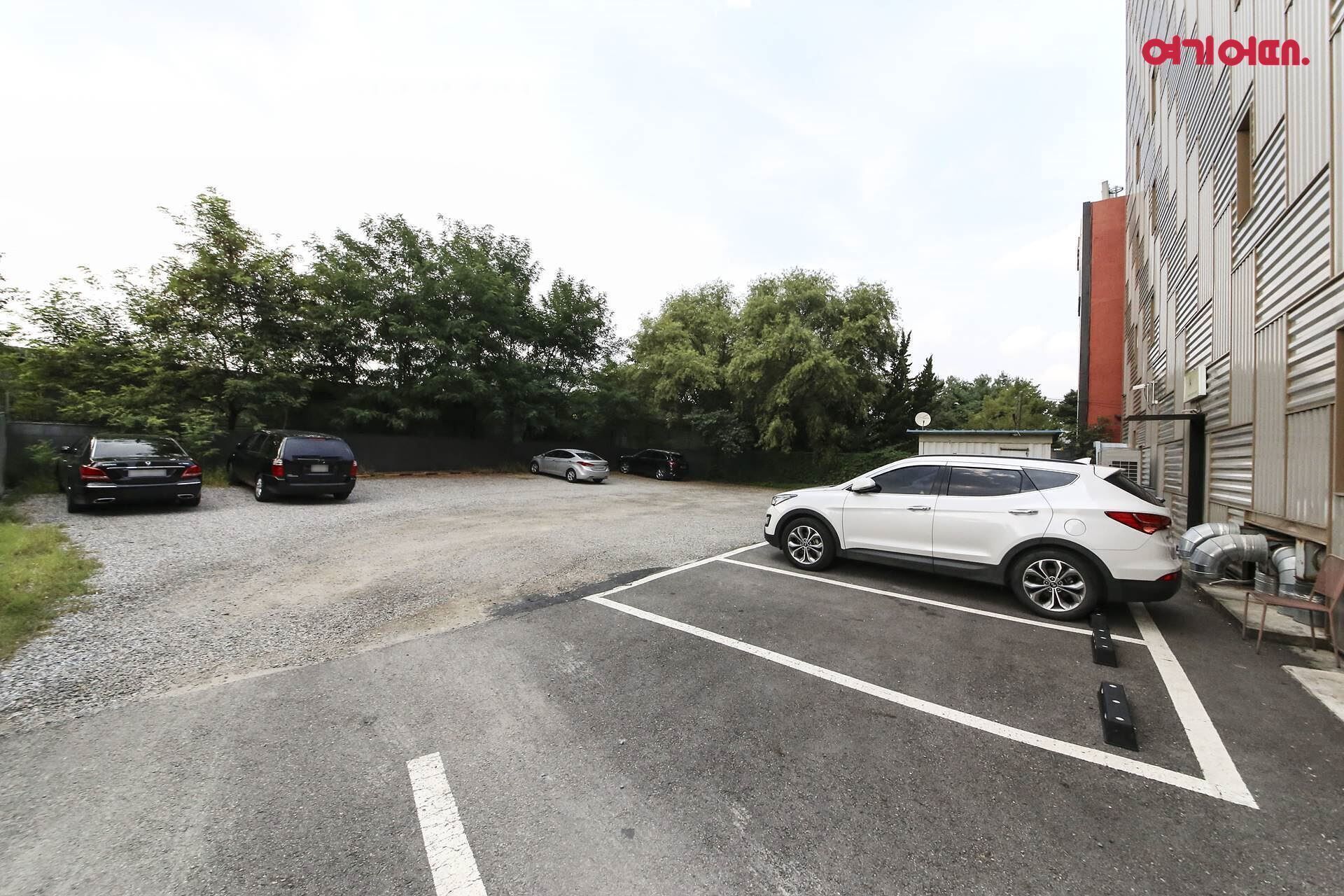 Uma área de estacionamento com vários carros, um edifício com fachada marcante e árvores ao fundo.