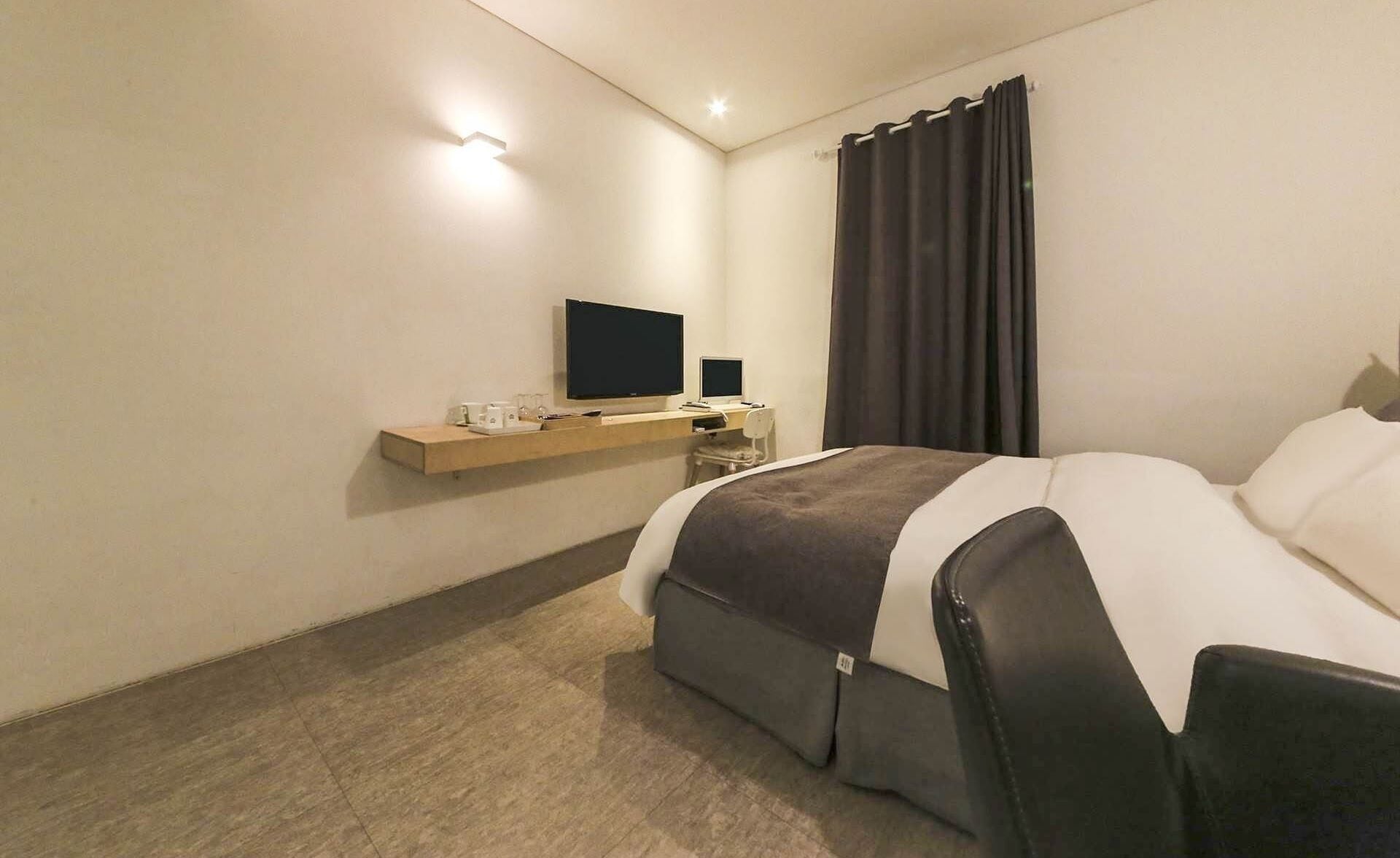 A modern hotel room with a bed, a desk with a computer, a flat-screen TV, and a small shelf with glasses.