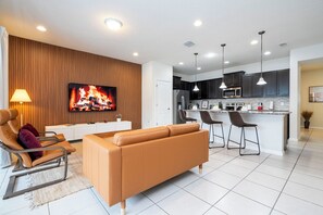 Un salon moderne avec un canapé, un téléviseur diffusant une image de cheminée et un coin cuisine doté de meubles sombres et d’un réfrigérateur en acier inoxydable.