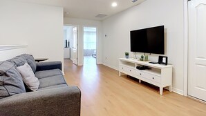 A modern living room with a grey sofa, a flat-screen TV on a white console, wooden flooring, and a door leading to another room.