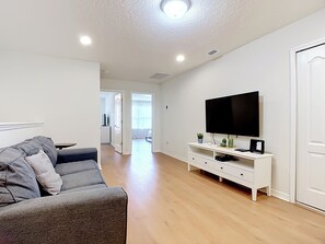 Un salon moderne avec un canapé gris, un téléviseur à écran plat posé sur une console blanche, un plancher en bois et une porte donnant sur une autre pièce.