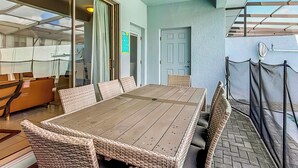 Une terrasse extérieure avec une table en bois et des chaises en osier, une porte vitrée donnant sur un espace intérieur, et une porte sur laquelle est affiché un panneau indiquant les règles de la piscine.