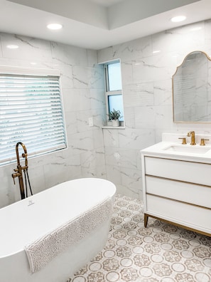 A modern bathroom with a freestanding bathtub, a vanity with gold fixtures, and a marble-tiled floor. - The Cozy Three-Bedroom House on Holly Lane (Bonita Springs)