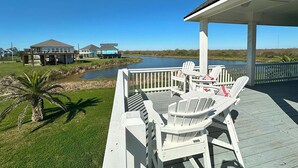 A wooden deck with white chairs and a table, overlooking a river and houses in the background.