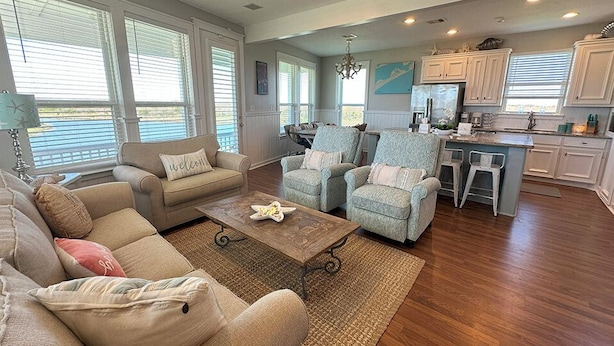A spacious living room with a sofa, armchairs, and a coffee table. There is a kitchen area with white cabinets and a dining table.