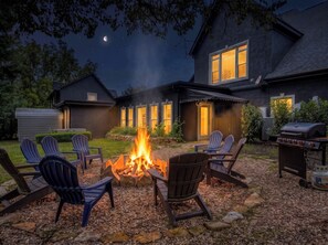 A backyard with a fire pit, Adirondack chairs, a grill, and a house with a dark exterior and lit windows.