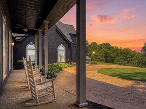 Ein überdachter Balkon mit Schaukelstühlen, ein Haus mit dunkler Fassade und Blick auf den Sonnenuntergang.
