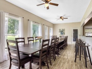 A dining area with a long wooden table, multiple chairs, ceiling fans, and large windows with curtains.