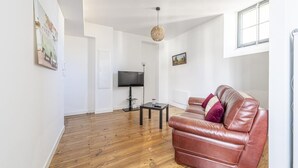 A spacious living room with a wooden floor, a leather sofa, a television, and a hanging light fixture. - Cœur de Pau - Parking included - L'Étoile (Pau)