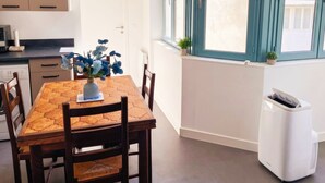 A dining area with a table, chairs, a vase with flowers, a window with curtains, and a white air conditioning unit. - Cœur de Pau - Parking included - L'Étoile (Pau)
