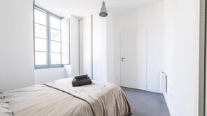 A minimalist bedroom with a large window, a bed with a beige blanket, and a white door. - Cœur de Pau - Parking included - L'Étoile (Pau)