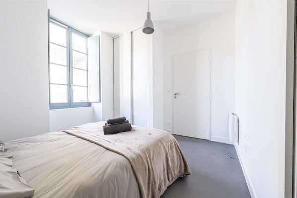 Et minimalistisk soveværelse med et stort vindue, en seng med et beige tæppe, og en hvid dør.