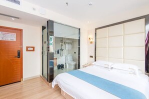 A hotel room with a bed, a wooden door, a glass partition, a bathroom, and a padded headboard.