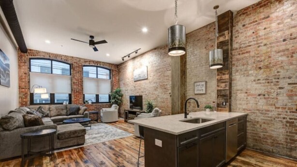 A spacious living room with a large sofa, a coffee table, and a TV. The room has a brick wall and wooden flooring. - Cozy Downtown Asheville Condo | Walk everywhere (Asheville)