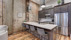 A modern kitchen with a large island, bar stools, stainless steel appliances, and exposed brick walls. - Cozy Downtown Asheville Condo | Walk everywhere (Asheville)
