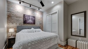 A bedroom with a bed, a nightstand, a dresser, a mirror, and a brick wall. - Cozy Downtown Asheville Condo | Walk everywhere (Asheville)