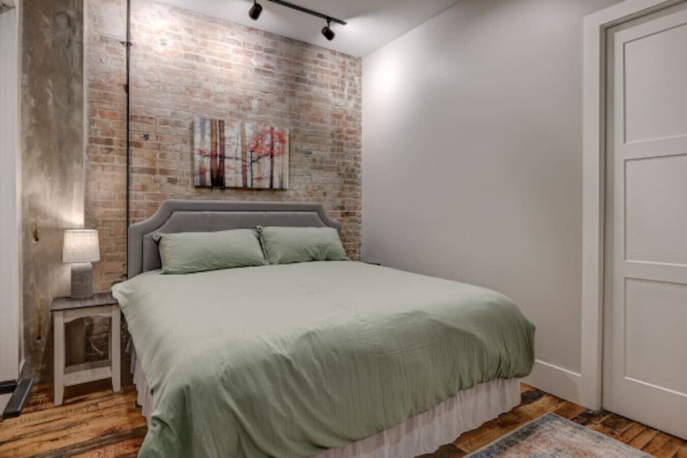 Un dormitorio con pared de ladrillo, una cama con cabecera gris, una mesita de noche con lámpara y una puerta.