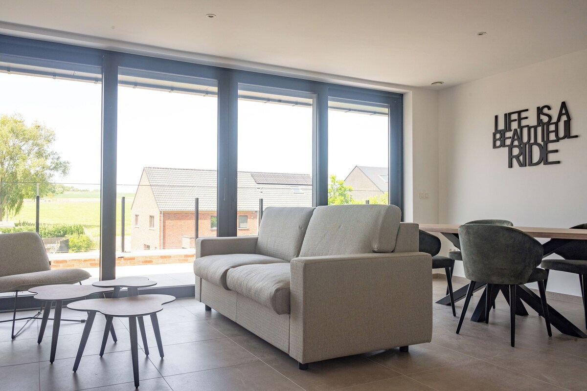 A modern living room with a sofa, two small tables, and a dining area with chairs. There is a large window providing a view of the outside.