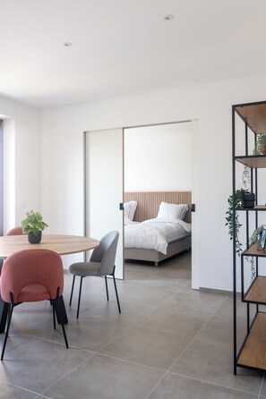 A modern hotel room with a wooden dining table, two chairs, a bed with a wooden headboard, and a black metal shelving unit with plants. - Flanders Cobblestone (Brakel)