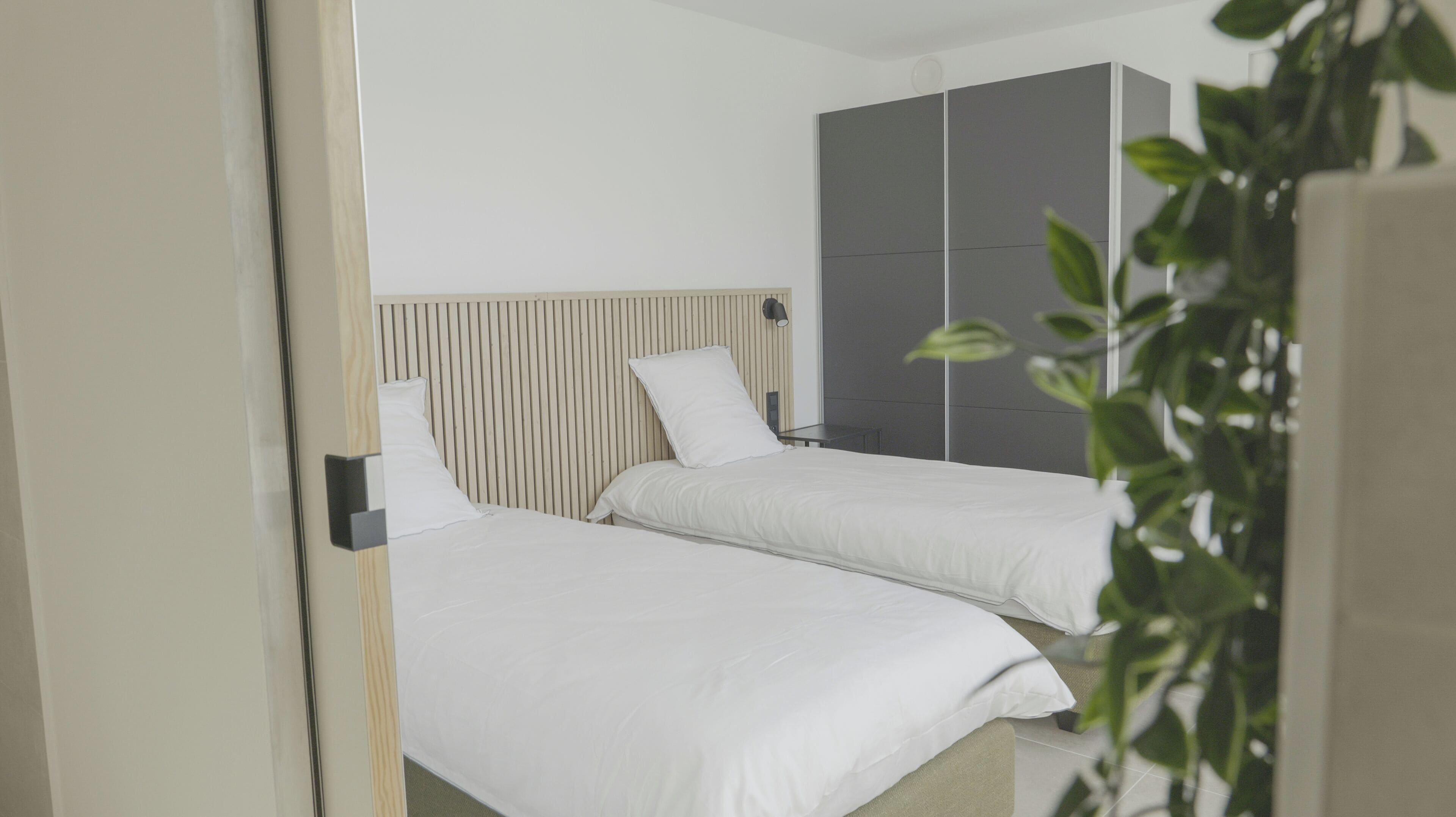A hotel room with two beds, a wooden headboard, a grey wardrobe, and a small table.
