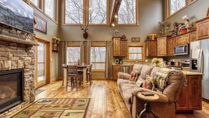 A cozy cabin interior with a stone fireplace, wooden floors, a dining area, a living room with a sofa and a coffee table, and a kitchen area.