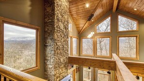 A high-ceilinged room with a stone fireplace, large windows, and a wooden staircase.
