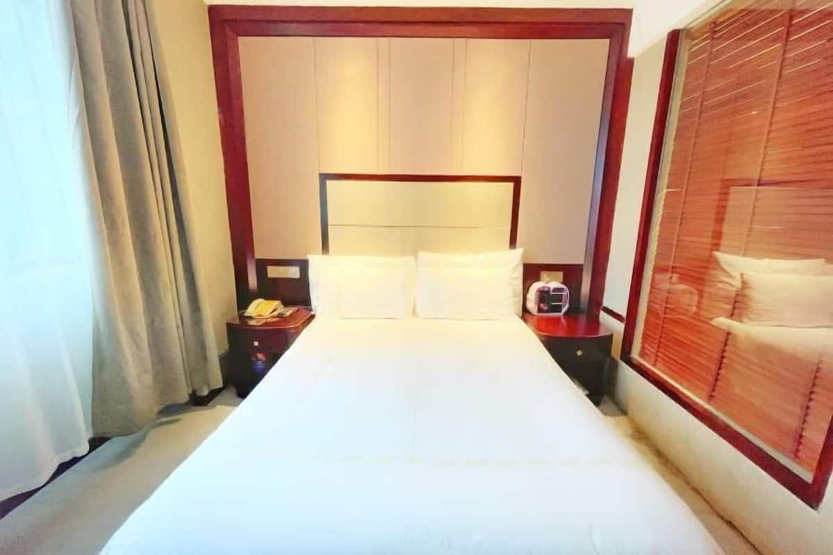 A hotel room with a large bed, a bedside table with a lamp, a telephone, and a window with blinds.
