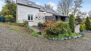 A house with a stone exterior, a patio with a table and chairs, and a garden with various plants and trees.