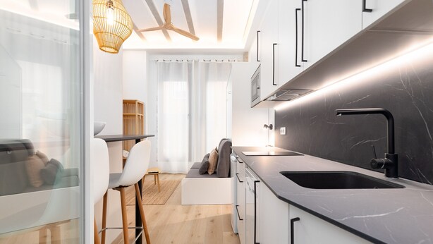 Een moderne keuken met een zwart aanrecht, witte kasten en een inbouwoven. Er is een eethoek met een witte tafel en stoelen, en een bank op de achtergrond.