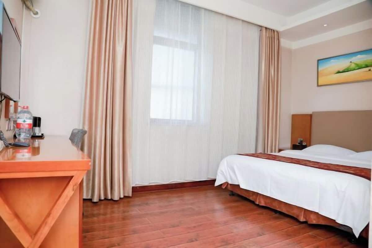 A hotel room with a wooden desk, a bed with white bedding, a painting on the wall, and a window with curtains.