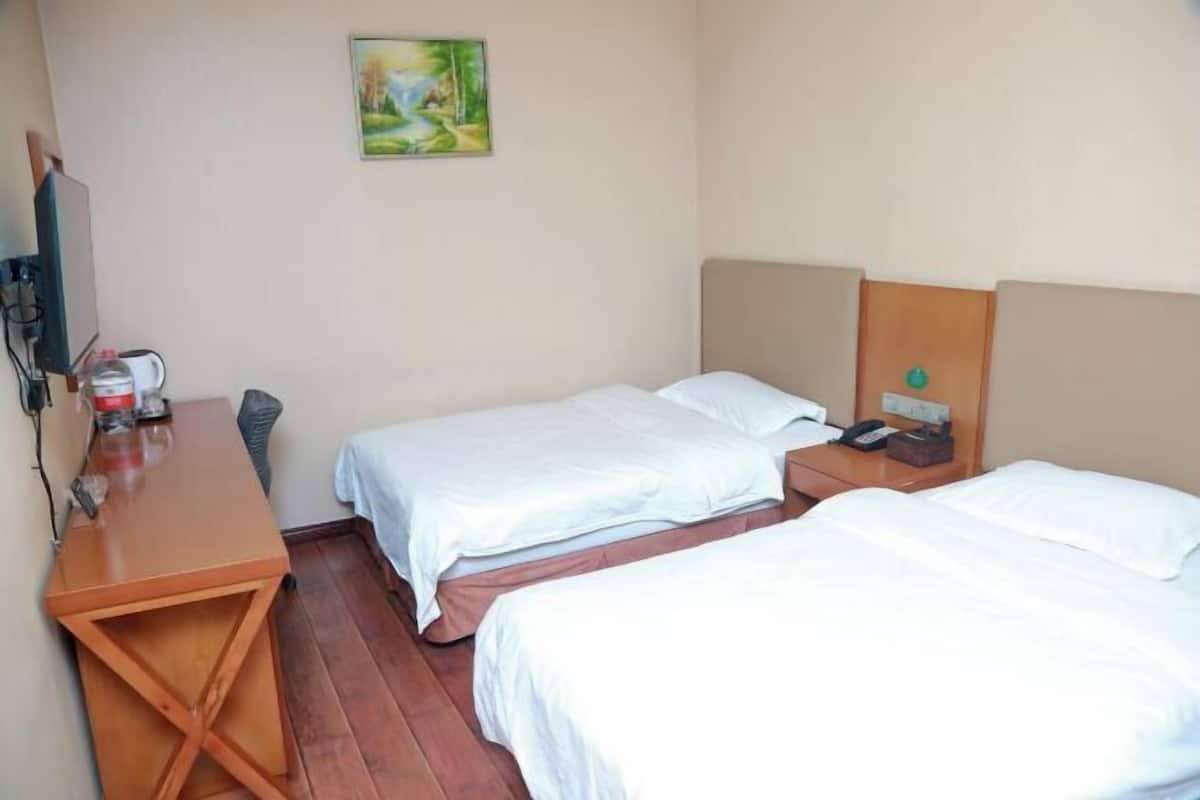 A hotel room with two beds, a desk, a chair, a painting on the wall, and a bedside table with a phone and a lamp.