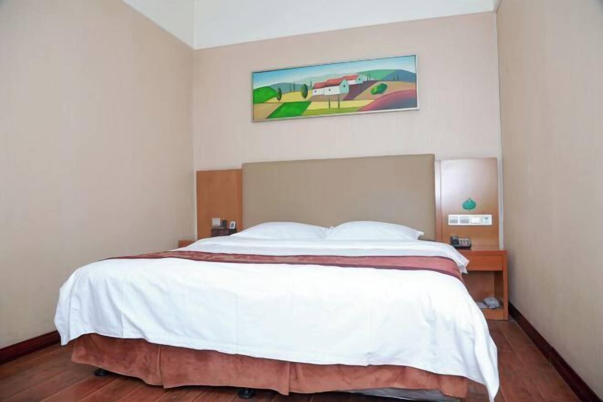 A hotel room with a bed, a painting on the wall, a nightstand, and a wall-mounted lamp.