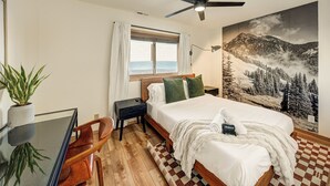 A hotel room with a bed, a desk with a chair, a ceiling fan, a window with curtains, and a mountain landscape painting. - Gorgeous Remodeled 3-Bedroom home in charming downtown Frisco with Private Sauna (Frisco)