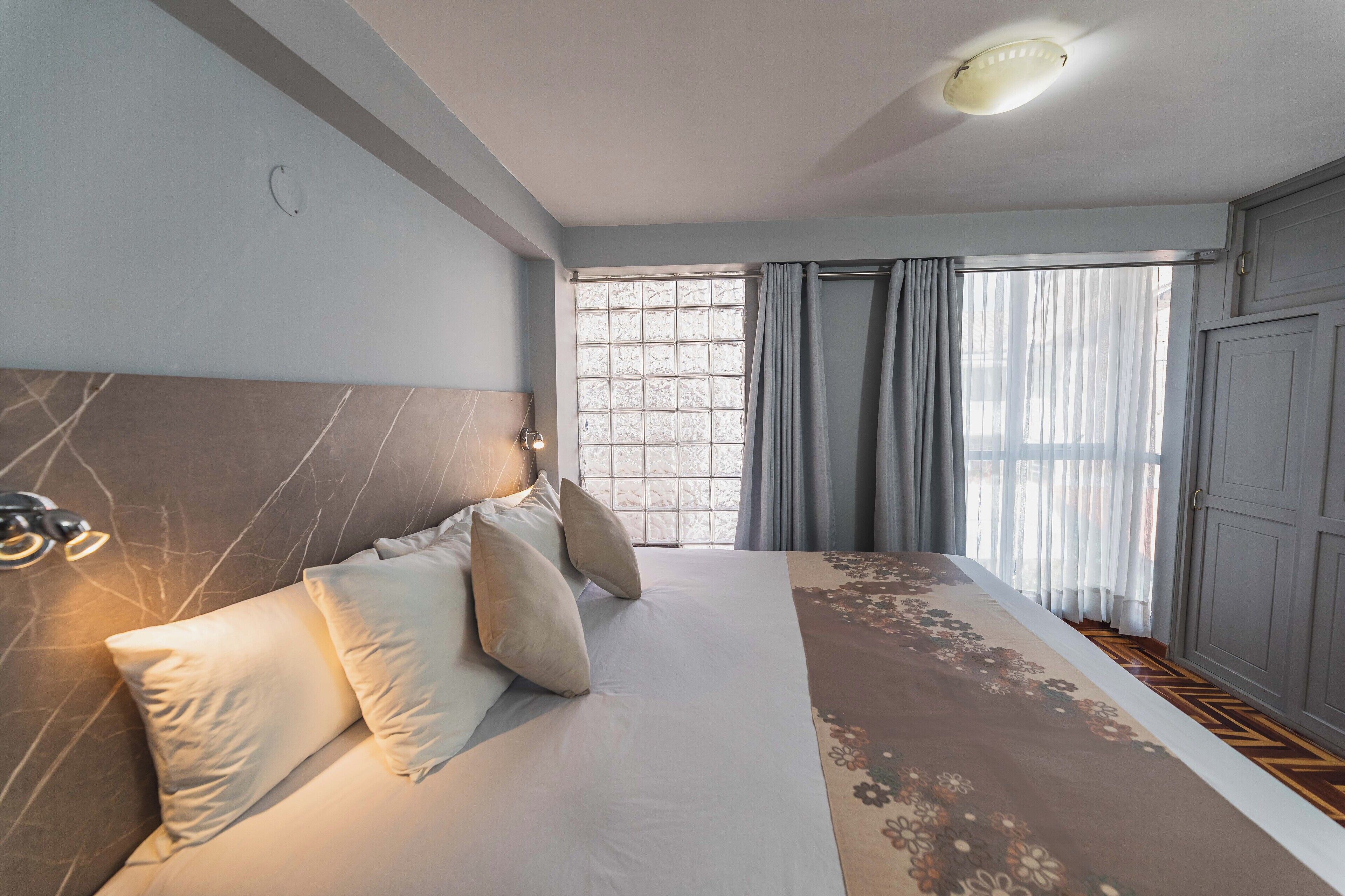 A bedroom with a large bed, a headboard with a light fixture, a window with curtains, and a wardrobe.